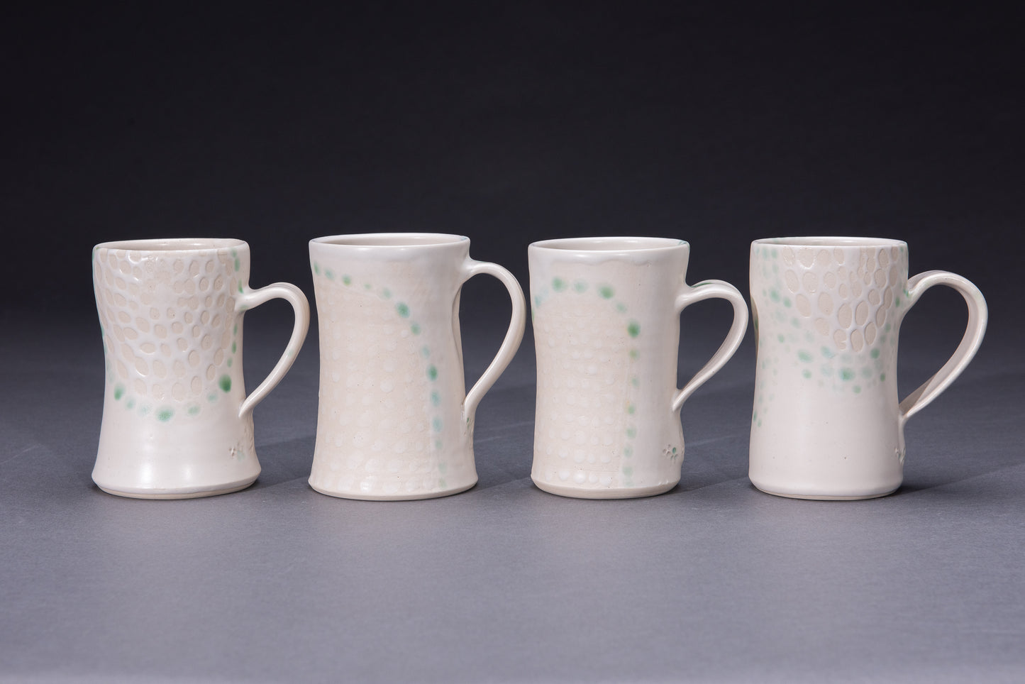 Four ceramic mugs featuring asmooth white glaze dotted with bare clay and green dot detail on a gray background