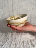Hand holding a ceramic bowl with brown and beige swirl pattern against a textured gray background