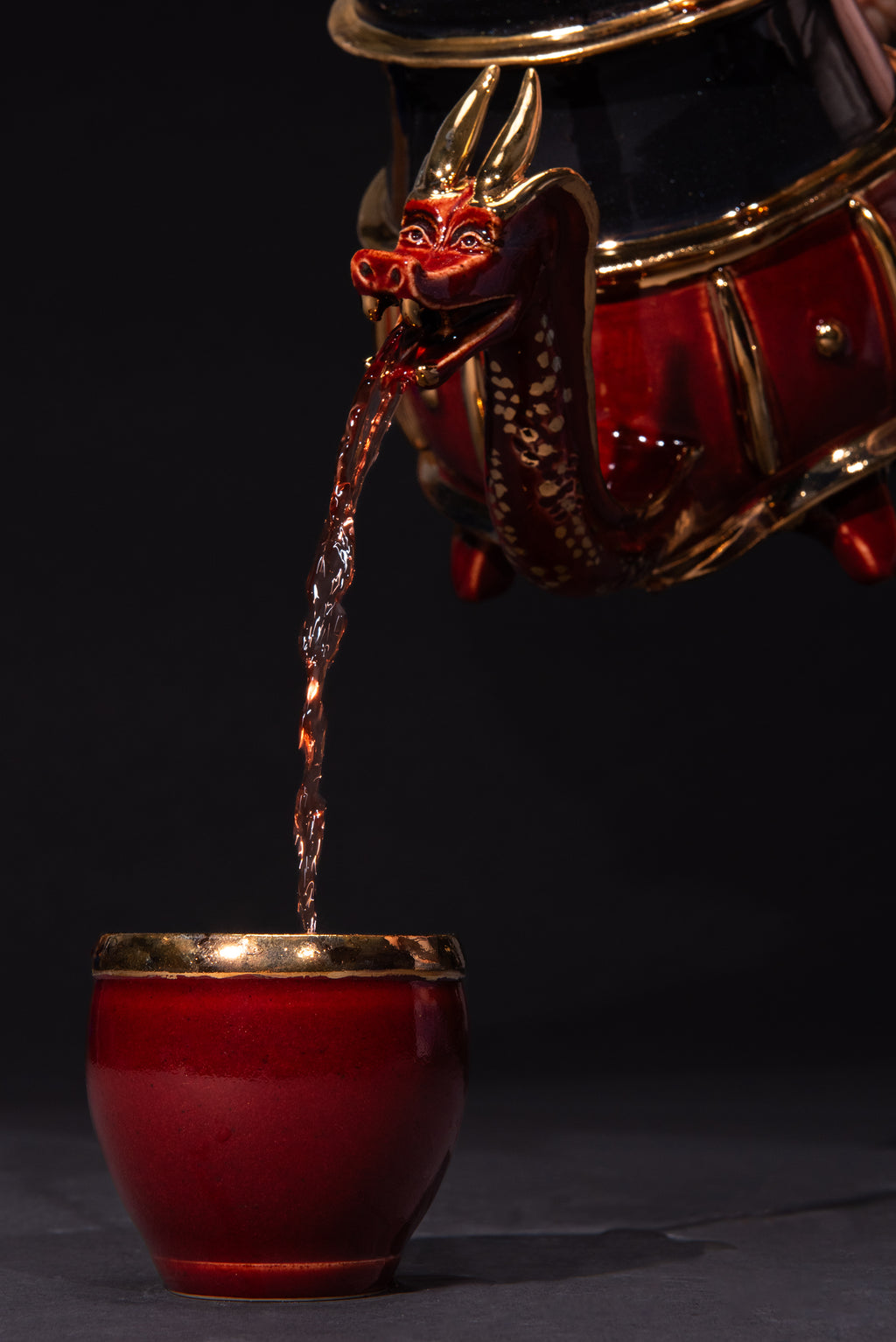 Red teapot with gold accents pouring liquid into a matching red cup on a black background