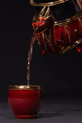 Red teapot with gold accents pouring liquid into a matching red cup on a black background