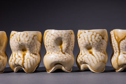 A set of sculptural ceramic drinking vessels in amber ash color, displayed in a row, showing an organic, uneven form with gentle indentations and an irregular rim.