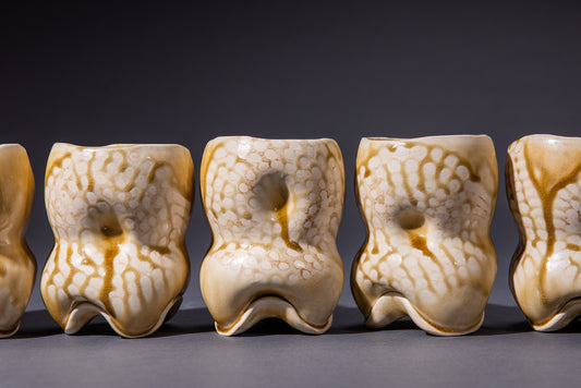 A set of sculptural ceramic drinking vessels in amber ash color, displayed in a row, showing an organic, uneven form with gentle indentations and an irregular rim.