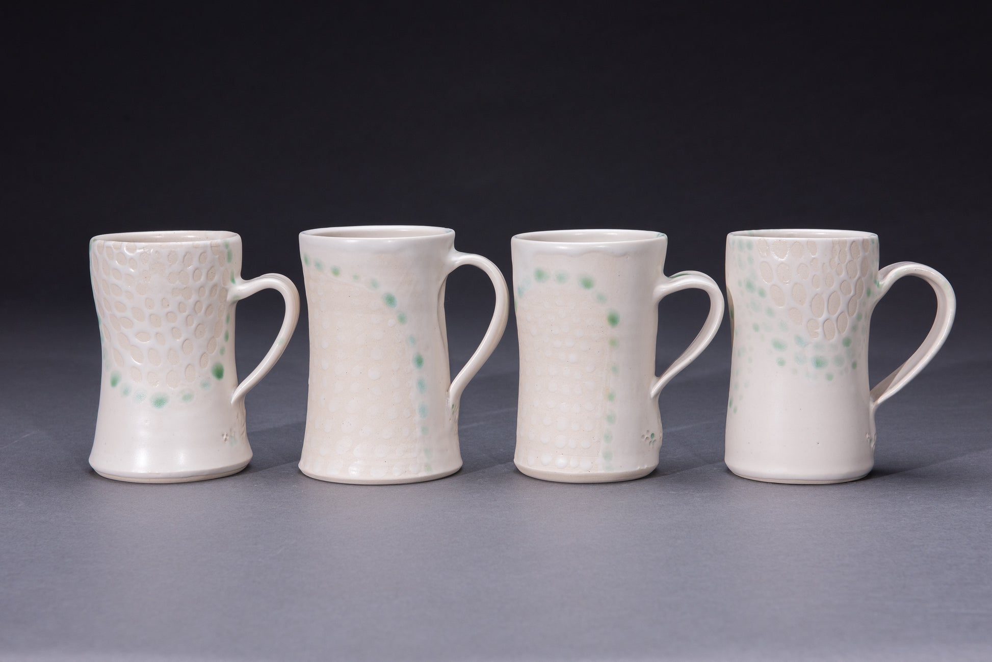 Four ceramic mugs featuring asmooth white glaze dotted with bare clay and green dot detail on a gray background
