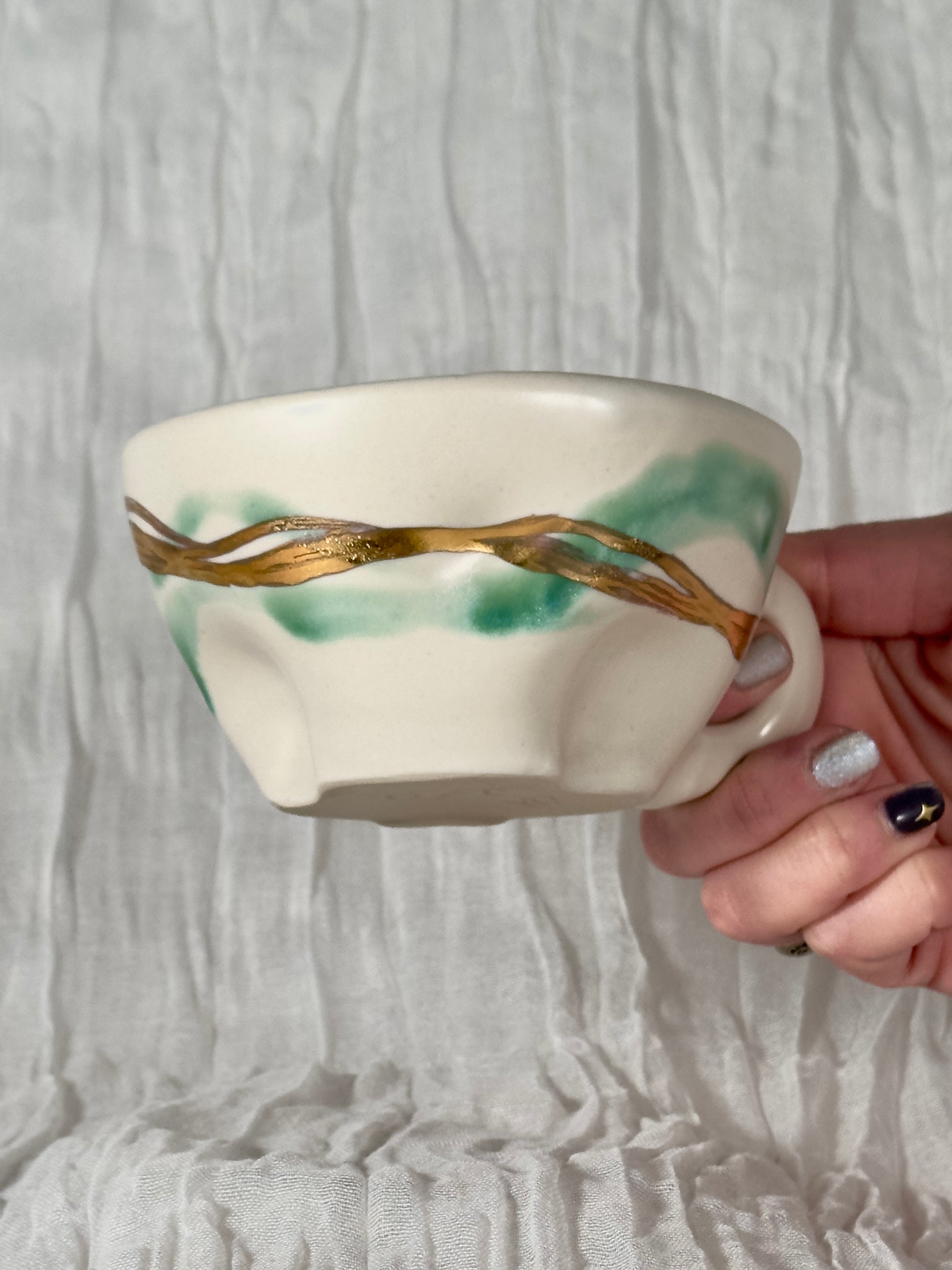 Hand holding a ceramic cup with green and gold design against a textured white background
